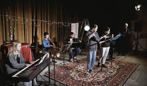 Eine jugendliche Band aus Keyboarderin, Cellist, Akkordeonist, zwei Flötistinnen und zwei Gitarissten (ein älterer). Alle stehen auf einem Teppich auf einer alternativ anmutenden Bühne.