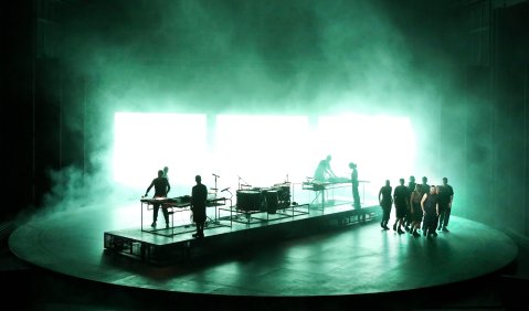 Die Bühne ist in grünen Nebel getaucht. Auf ihr steht ein mehrere Meter großes, kreisrundes Podest. In der Mitte die aufgebauten Percussion-Instrumente, rechts der Chor. Dahinter drei große Lichtpanele.