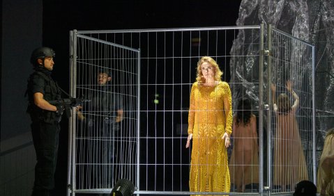 Médée im gelben Kleid inmitten eines Bauzaun-Quadrats bewacht von Soldaten.