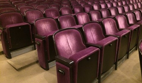 Sehr violett-weinrote Satinbezogene Theaterbestuhlung. Beiger Teppichboden darunter.
