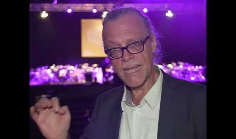 Rainer Nonnenmann, ein Mann mit zurückgebundenem silbernen Haar, Brille und Bart, steht in einem Saal der lila erleuchtet ist. im Hintergrund sieht man das gehende Publikum und die Drumsets die abgebaut werden.