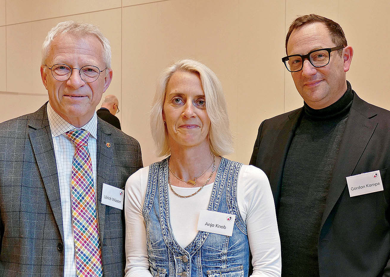 Zwei Männer in Anzügen und eine Frau in einer Jeansweste stehen mit angesteckten Namensschildern für ein Foto zusammen.