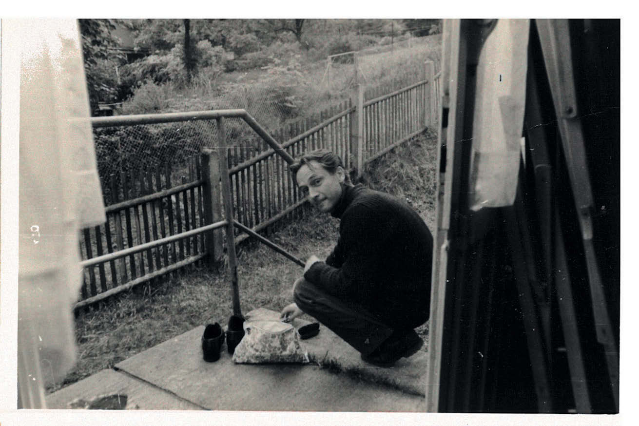 Schwarzweißbild eines Mannes mit kurzen schwarzen Haaren, der auf Treppenstufen vor einem Einfamilienhaus Schuhe putzt.