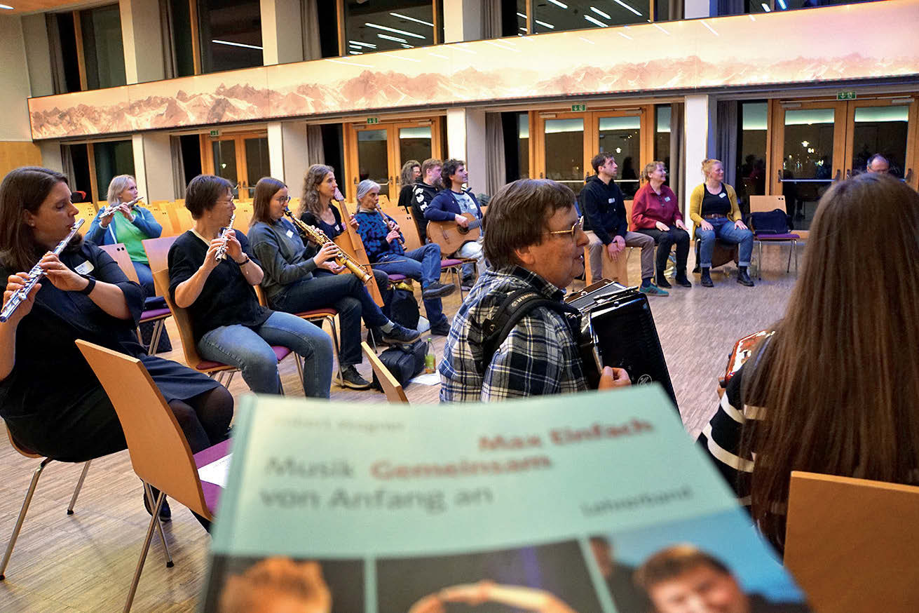 Eine Gruppe Erwachsener Menschen sitzen mit verschiedenen Instrumenten in einem großen Halbkreis. Am unteren Ende des Bildes wird ein verschwommener Buchtitel von "Max Einfach. Musik gemeinsam von Anfang an" ins Bild gehalten.