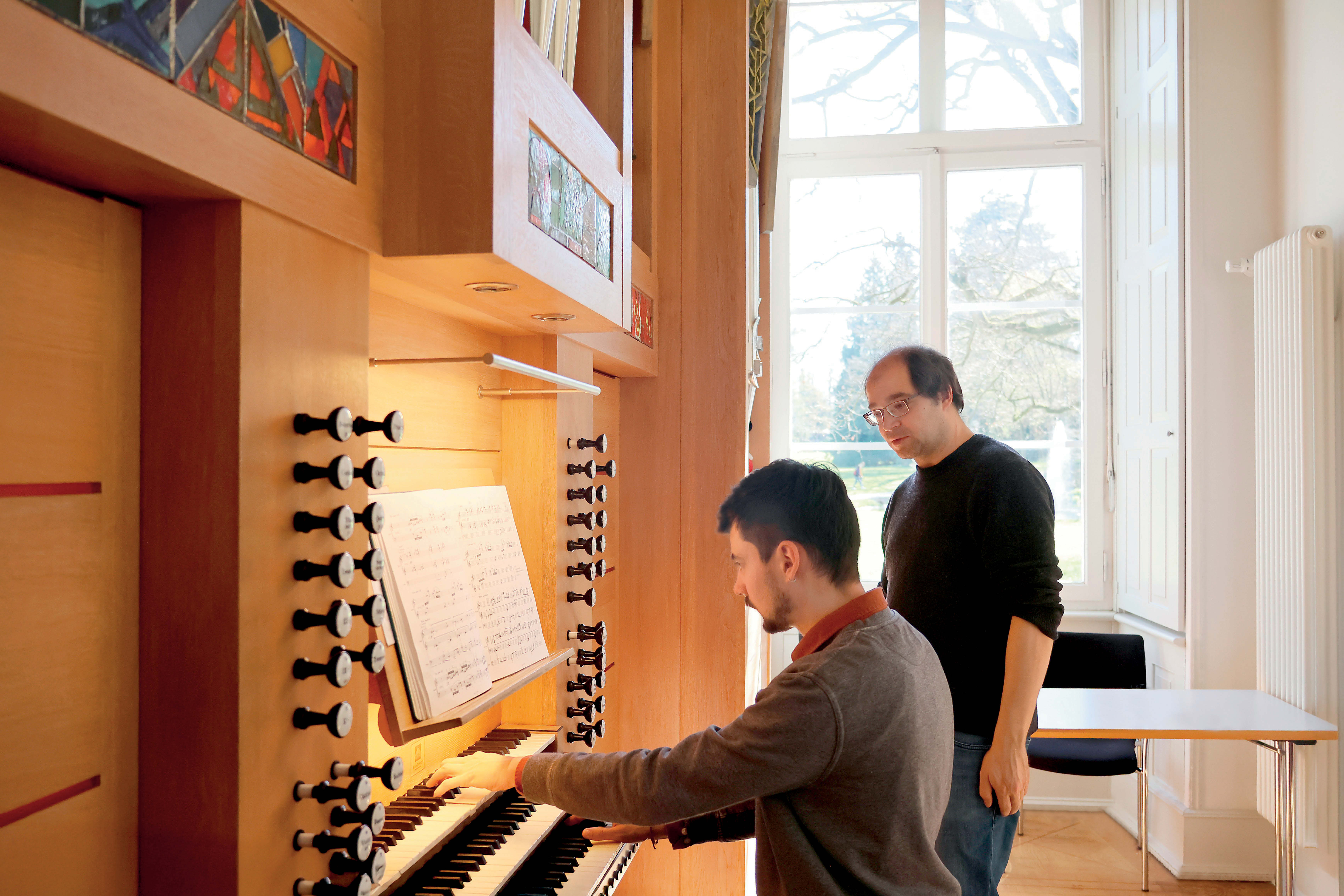 Orgelunterricht an einem Sonnenlichtgefluteten Orgelstuhl: Ein jüngerer Mann sitzt an einer Dreimanualigen Orgel. Ein etwas älterer Mann beobachtet ihn dabei. Im Hintergrund ist ein großes Fenster mit klarem Glas.
