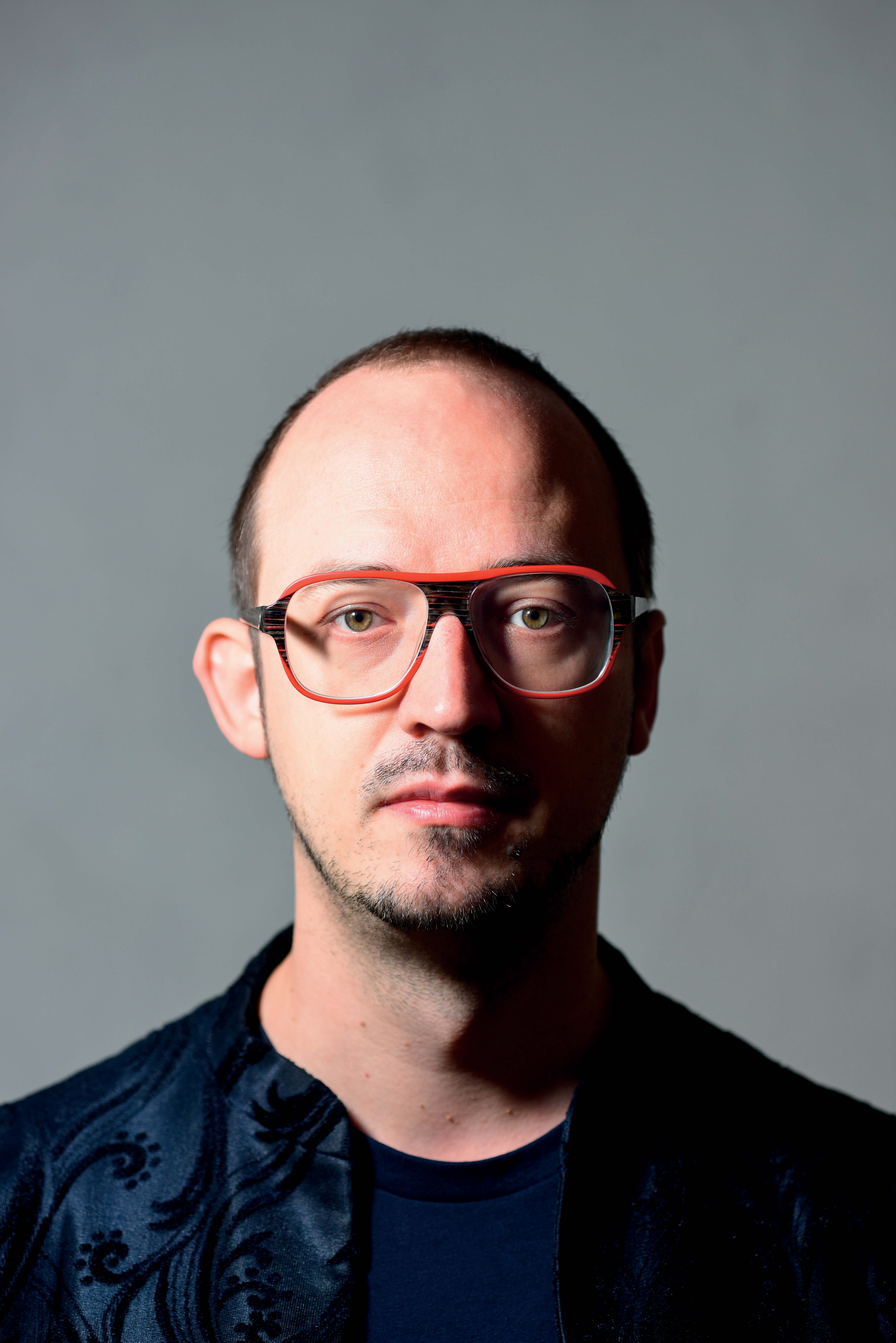 Ein Mann von unklarem Alter, kurzen schwarzen Haaren, leichtem Bartwuchs und einer großen Halbrunden roten Brille. Er steht vor einem grauen Hintergrund und schaut neutral in die Kamera.