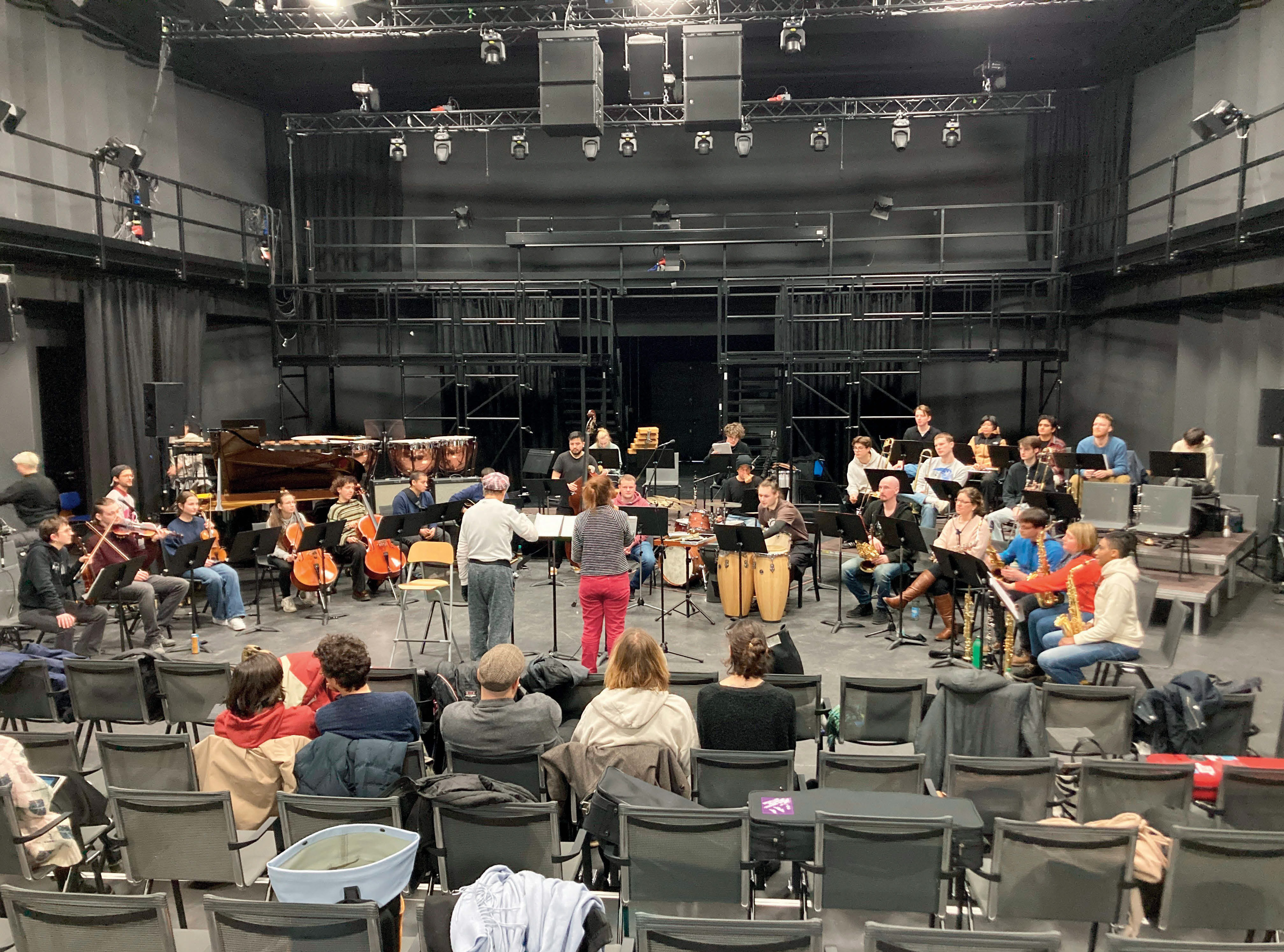 Eine rohe Theaterbühne (ohne Bühnenbild und mit viel sichtbarer Technik) auf der ein großes Ensemble probt. Im Zuschauerraum sitzen einzelne Menschen.