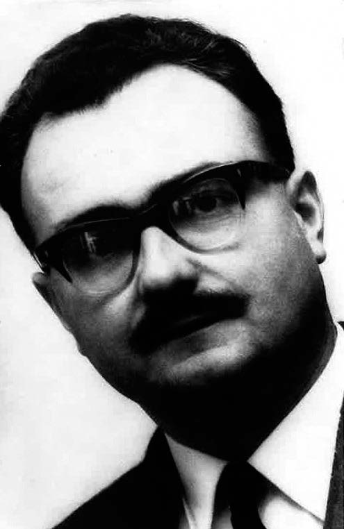 Unscharfes Schwarzweiß-Porträt eines Mannes in Anzug mit Hornbrille, Oberlippenbart und kurzen schwarzen Haaren.