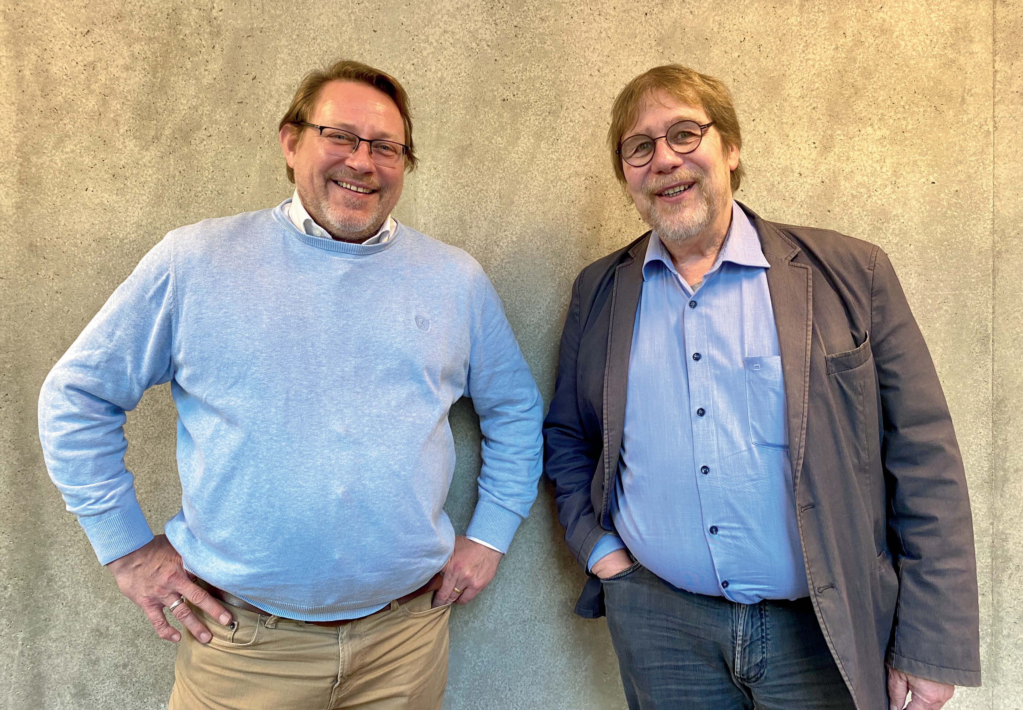 Zwei Herren im höheren mittleren Alter mit blauen Hemden (einer mit Sakko), kurzen Haaren und Brille stehen vor einer Beigen Wand und lächeln freundlich für das Foto.