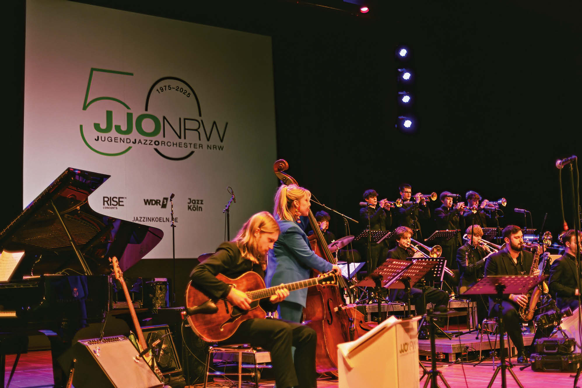 Bigband mit Fokus auf Bass-/Rhythmussection und einem Hintergrundtransparent "50 JJONRW"