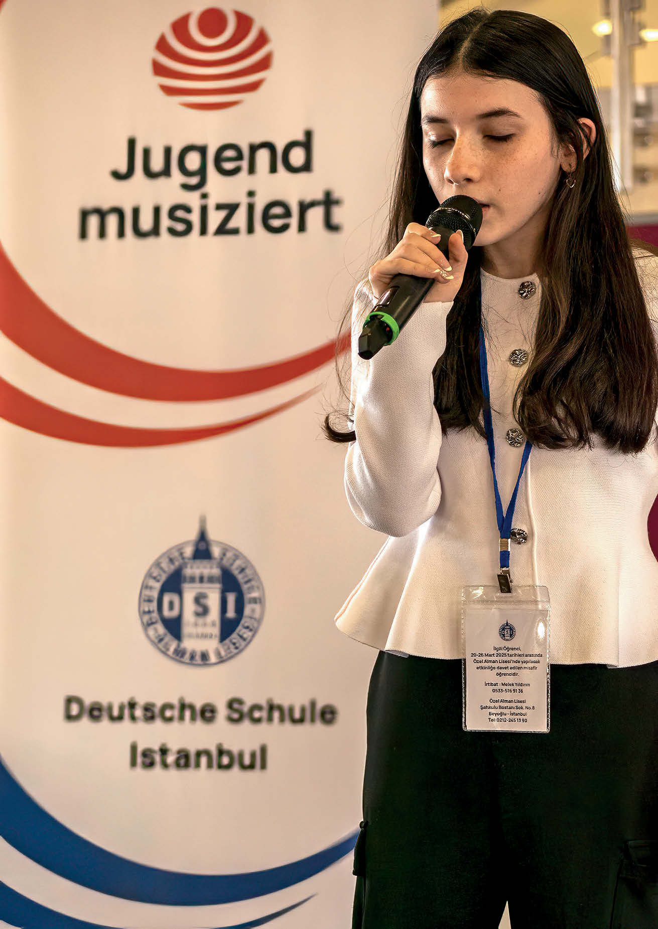 Ein Mädchen in schwarzweißen Klamotten mit langen schwarzen Haaren singt in ein Mikrofon. Hinter ihr ein Transparent: "Jugend musiziert / Deutsche Schule Istanbul"