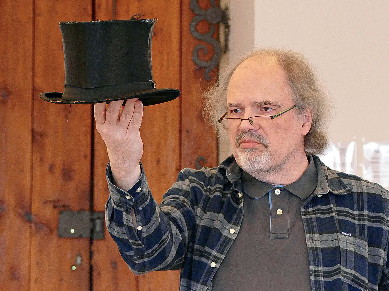 Ein Mann mit längeren, grauen Haaren, hoher Stirn, Brille und grau kariertem Hemd hält einen Zylinder vor sich und betrachten ihn eingehend.