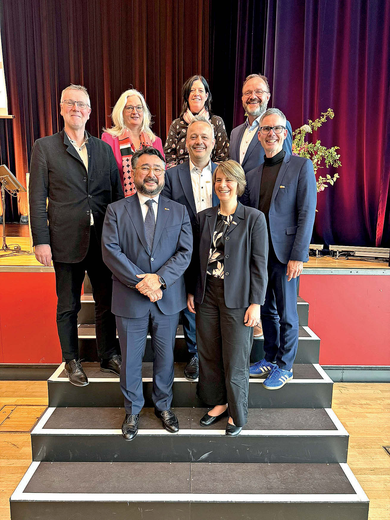 Auf einer Treppe stehen mehrere Erwachsene zu einem Gruppenfoto zusammen. Sie tragen meist Anzüge etc.