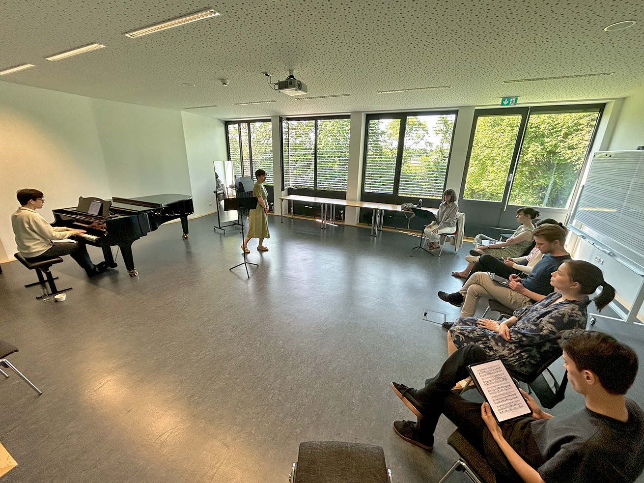 Ein Seminar mit Flügel und einer Sängerin. Mehrere Teilnehmer:innen sitzen als Publikum und Teilnehmer zusammen.
