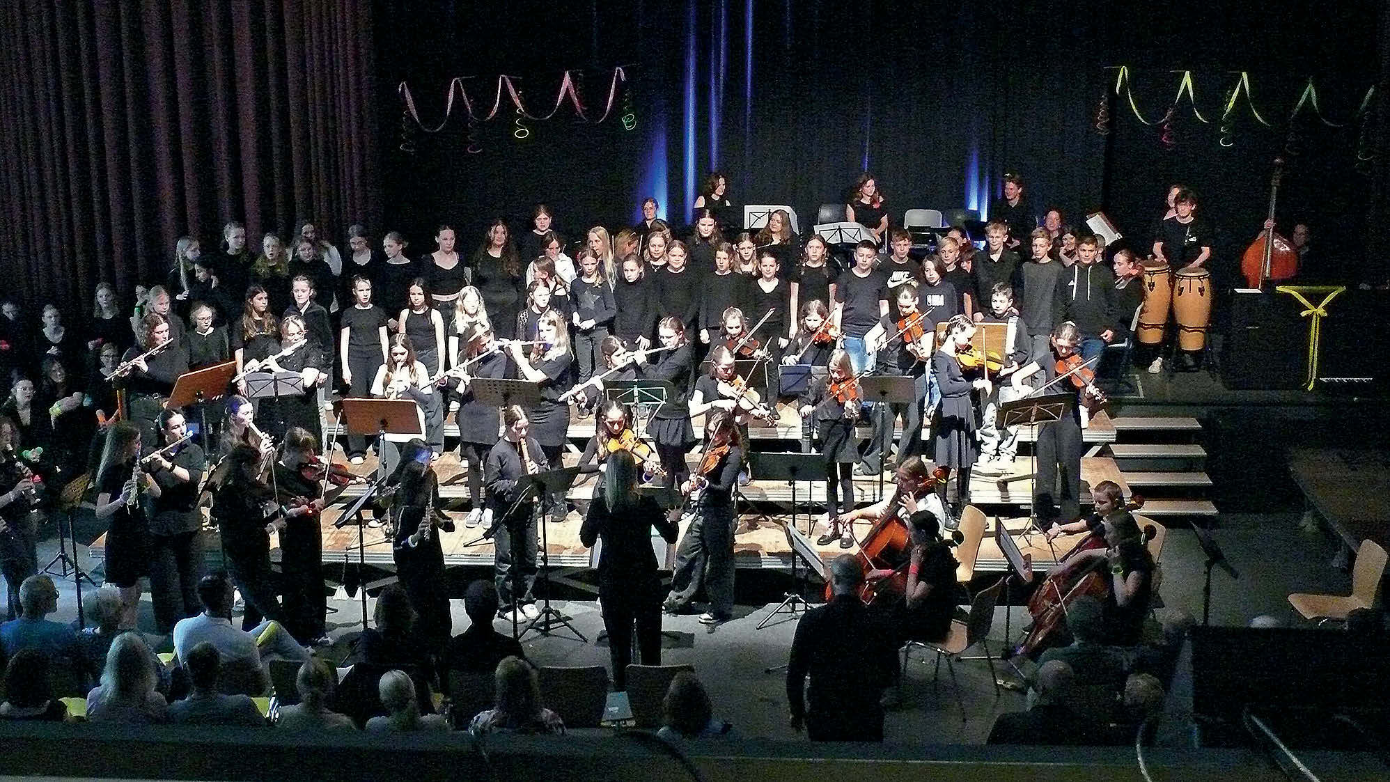 Eine große dunkle Bühne einer Mehrzweckhalle oder Aula die mit sehr vielen Kindern und Jugendlichen gefüllt ist. Die meisten tragen Schwarz und spielen Orchesterinstrumente.