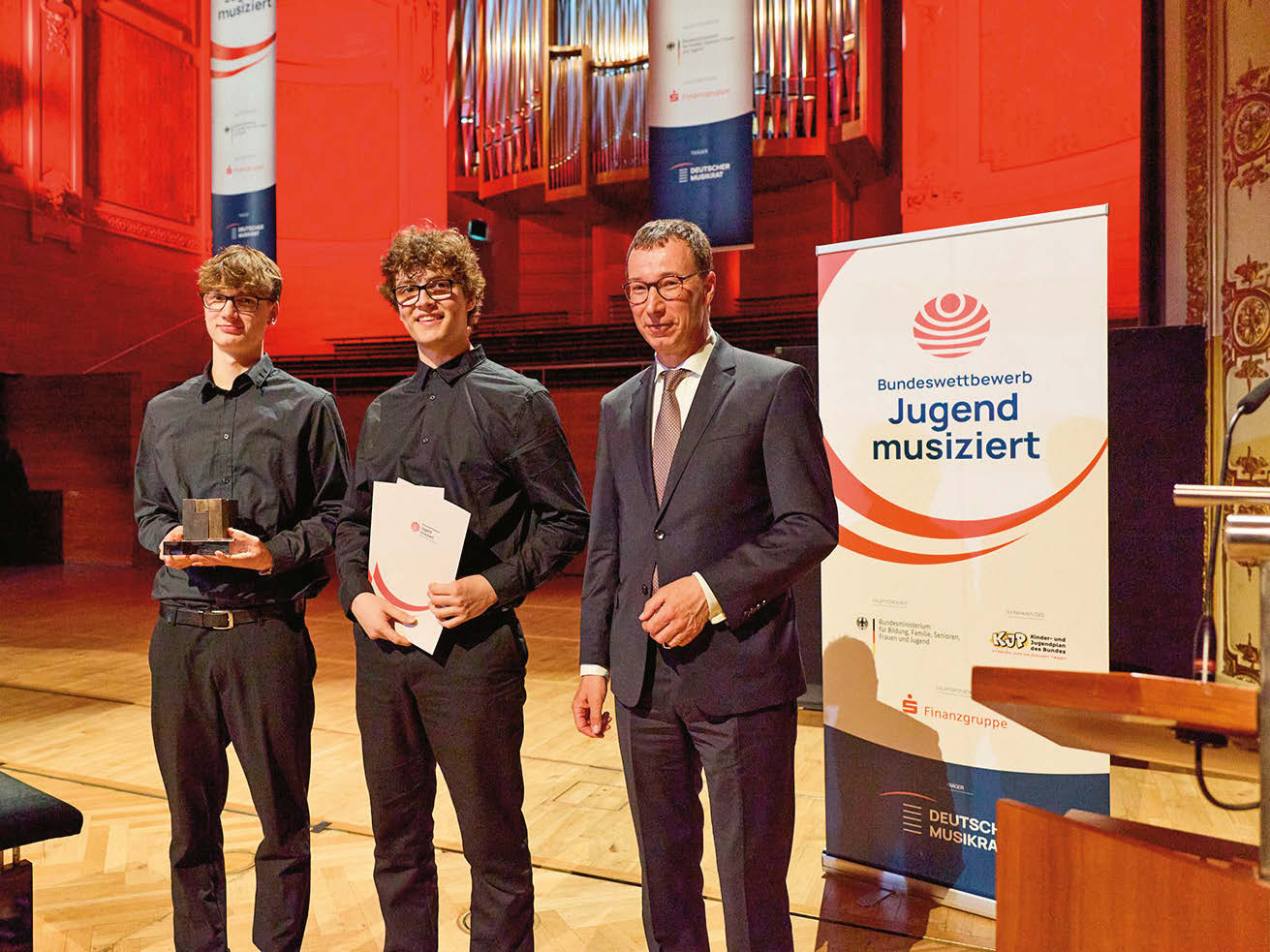 Zwei ältere Jugendliche (schwarz gekleidet) und ein Mann (im Anzug) stehen mit Urkunden und einem kleinen Metall-Preis in einem Konzertsaal. Im Hintergrund tront das Pfeifen-Bouquet einer Orgel.
