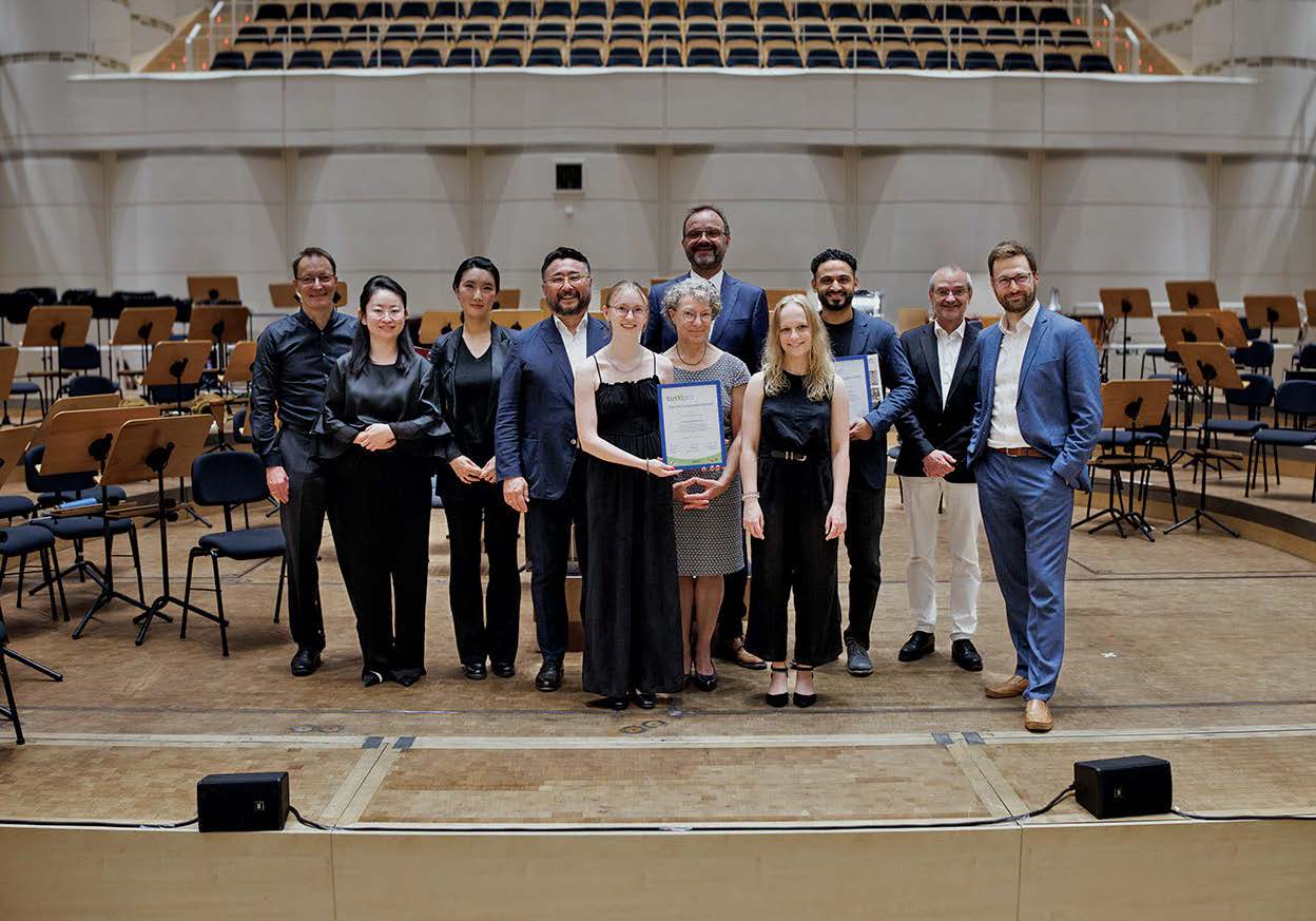 Mehrere Menschen stehen in dem Konzertsaal in dem im Beitragsbild das Orchester aufgetreten ist in schicken Klamotten zu einem Gruppenbild zusammen. In der Mitte halten zwei Frauen ein Dokument.