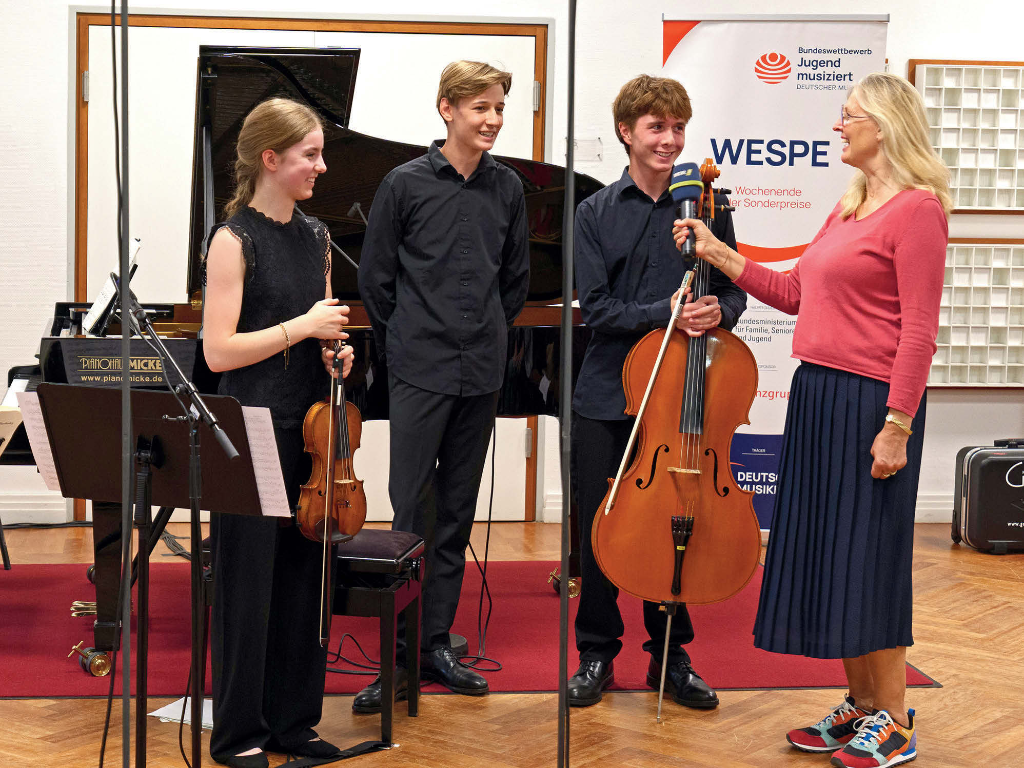 Eine Moderatorin vom WDR (Pinker Pullover, blauer Rock, blonde Haare) interviewt den Cellisten eines Klaviertrios. Die anderen (Violinistin und Pianist) aus dem Trio stehen neben ihm.