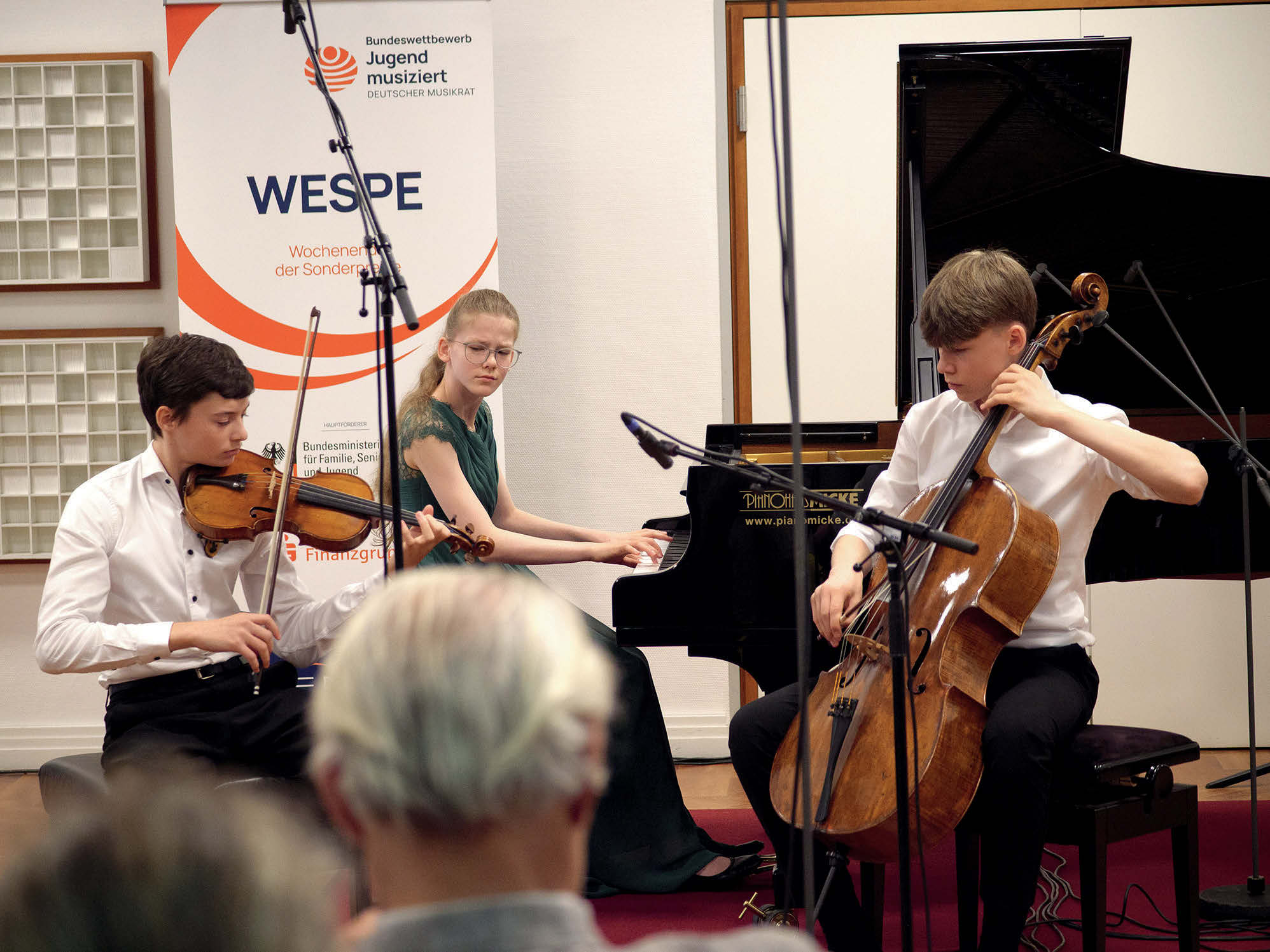 Ein Klaviertrio (die Streicher zwei Jungen, am Flügel ein Mädchen) spielen ein Konzert vor einem WESPE-Aufsteller. Man sieht Mikrofone: Der Auftritt wird mitgeschnitten.