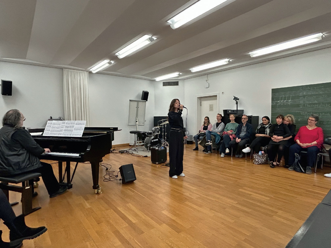Eine S&auml;ngerin mit Mikrofon (lange schwarze Kleidung, wei&szlig;e Schuhe) in einem kleineren Musikunterrichtsraum. Vor der Tafel an der Wand zu ihrer linken sitzen einige Zuh&ouml;rer:innen an der Wand. Zu ihrer Rechten steht ein Fl&uuml;gel mit einem erwachsenen Pianisten.