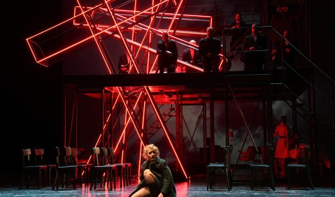 Eine junge Frau kauert auf der Bühnenmitte. Vor dem schwarzen Hintergrund stehen 3 verschieden geneigte Kreuze aus roten Leuchtstoffröhren. Von höheren Ebenen schauen Männer auf die Frau herab.