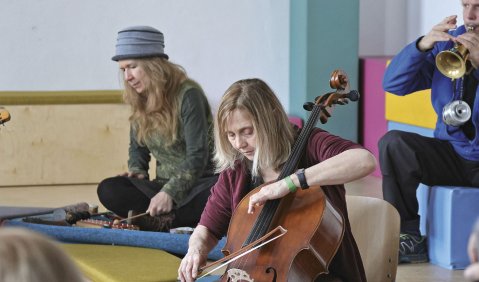 In einer Turnhalle spielt eine Frau auf dem Boden Glockenspiel, eine andere im Vordergrund Cello und im Hintergrund spielt ein Trompeter. An seiner Trompete hängt ein Dämpfer herunter.