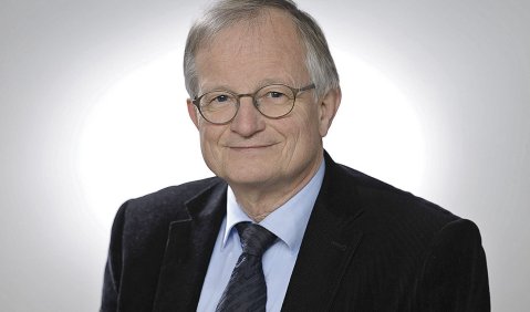Portrait eines lächelnden Herrn mit silbirg-grauen, kurzen Haaren und Brille in einem dunklen Anzug mit Krawatte