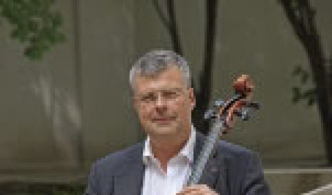 Christian Höppner sitzt draußen, im lockeren Anzug ohne Krawatte, hinter seinem Cello.