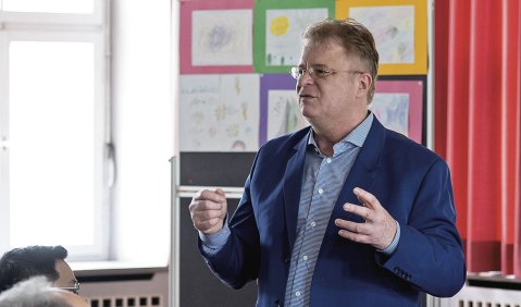 Gestikulierend steht von Pragenau mit kurzen Haaren, Brille und Sakko vor dem Publikum. Eine bunte Tafel im Hintergrund.