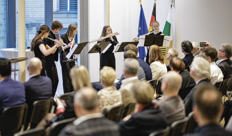Fünf schwarz gekleidete Flötistinnen spielen vor einem Kongresspublikum. Im Hintergrund die Europäische, Deutsche und Sächsische Flagge.