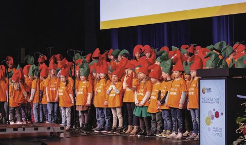 Ein großer Kinderchor mit grünen und roten Zwergenmützen und orangenen T-Shirts steht auf einer Bühne zum singen bereit.
