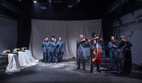 Eine kleine Bühne, abgesteckt mit weißen hängenden Laken, mit zwei Gruppen von Menschen mit blaugrauen Overalls. Weiter hinten ohne und weiter vorne mit Instrumenten. Links stehen drei Nähtische mit Maschine und weißen Laken.