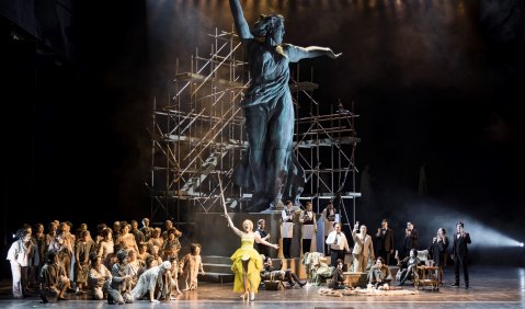 Eine riesige Statue mit Baugerüst drumherum. Über ihr sieht man nur noch die Theaterbeleuchtung. Zu ihren Füßen tummeln sich allerlei Menschen. Eine Frau in gelben Kleidern ahmt die Pose der Statue nach: Der rechte Arm mit Schwert nach oben gereckt, der zweite gerade nach hinten gestreckt.