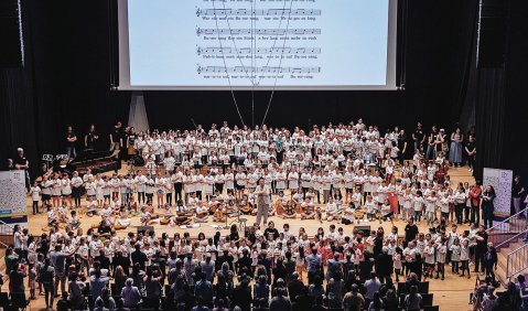 Auf der Bühne unter einer Leinwand mit angestrahlten Noten haben sich alle Beteiligten zum gemeinsamen Musizieren versammelt.