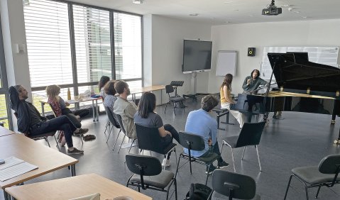 Zwei Menschen sitzen am Klavier, eine Gruppe hört in einem Seminar-Setting zu.