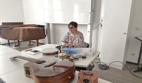 Die Komponistin sitzt an einem Tisch mit diversen kleinen Instrumenten. Sie trägt eine Brille, eine bunte Bluse und hat die Hände auf einem Gerät. Wahrscheinlich ein Gerät zur Wiedergabe von Blindenschrift.