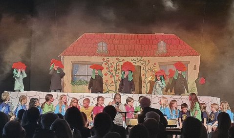 Das selbstgemachte Bühnenbild zeigt das Bild eines Hauses. Davor darstellende Kinder als Blumen oder Pilze mit roten Kopfbedeckungen.