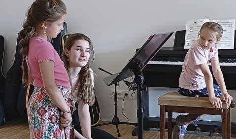 Eine Sudentin unterrichtet zwei Schülerinnen: Sie hockt neben einer stehenden, Geigenschülerin, daneben sitz eine Schülerin am E-Piano und dreht sich zu den beiden.