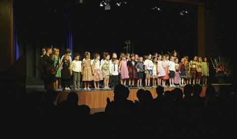 Ein bunter Kinderchor steht gut beleuchtet im sonst dunklen Saal