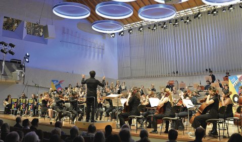 Eine grauer Konzertsaal, über dem Orchester hängen runde, blaue Klangreflektoren.