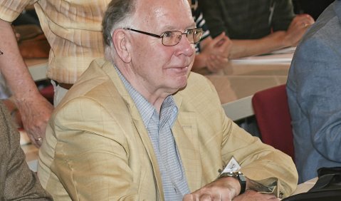Ein älterer Mann mit Brille und beigem Sakko auf einer Tagung.