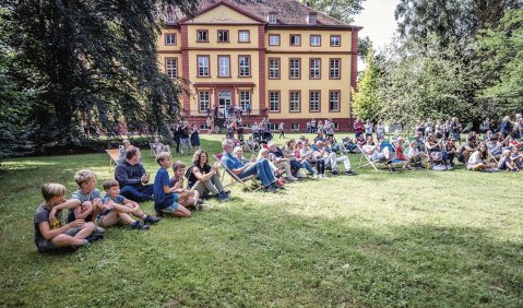 Ein altehrwürdiges Gebäude, davor ein großer Garten mit lauter Kindern, Jugendlichen und Erwachsenen die wohl einen nicht im Bild befindlichen Auftritt verfolgen.