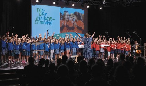 Eine Bühne in einer dunklen Halle. Im Hintergrund eine Projektion mit dem Titel der Veranstaltung. Davor auf der Bühne ein Kinderchor: Rechts Kinder in roten, links in blauen T-Shirts.