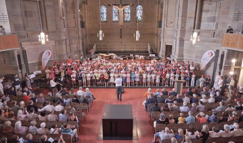 Eine volle Kirche. Auf der Bühne ein riesiger bunter Kinderchor.