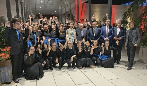 Die Orchestermitglieder in schwarz mit blauem Accessoire jubeln (teils zurückhaltend) neben den genannten Personen in die Kamera.