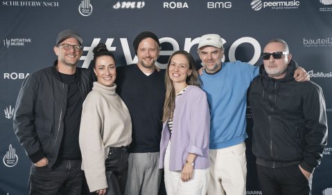 Eine bunte Truppe von Popmusikerinnen und Popmusikern vor einer Fotowand mit Sponsoren