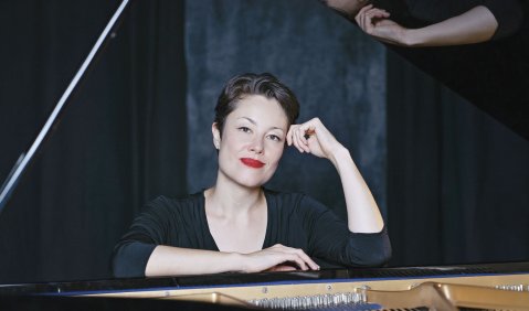 Pianistin mit Kurzhaarfrisur und stark geschminkten Lippen. Schaut aufs Klavieraufgestützt in die Kamera.
