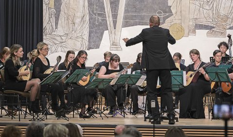 Ein Madolinenorchester während des Auftritts. Hauptsächlich Frauen spielen an grünen Noten sitzend in schwarz gekleidet.