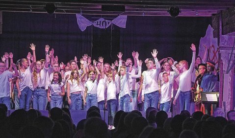 Ein Jugend- bzw. Kinderchor mit blauen Jeanshosen und weißen Oberteilen winkt von der Bühne.