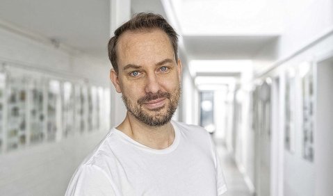 Ein jüngerer (mittelalter) Mann mit dunklem kurzen Bart, dunklen kurzen Haaren, weißem T-shirt in einer weißen Galerie